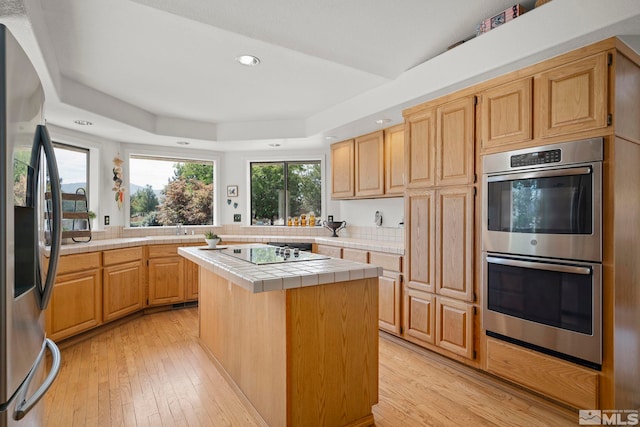 kitchen with light wood-type flooring, tile counters, light brown cabinetry, a kitchen island, and appliances with stainless steel finishes