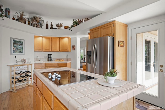 kitchen with light wood-type flooring, black electric stovetop, stainless steel fridge, tile countertops, and a center island