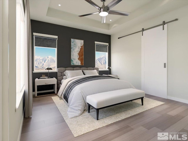 bedroom featuring a tray ceiling, light hardwood / wood-style flooring, ceiling fan, and a barn door