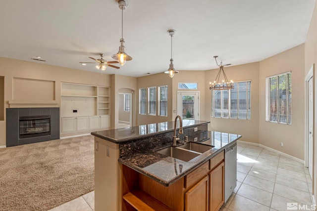 kitchen with a tiled fireplace, ceiling fan with notable chandelier, sink, dark stone counters, and a center island with sink