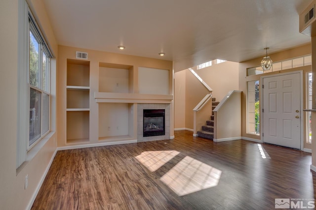 unfurnished living room with built in features, dark hardwood / wood-style floors, a tiled fireplace, and an inviting chandelier