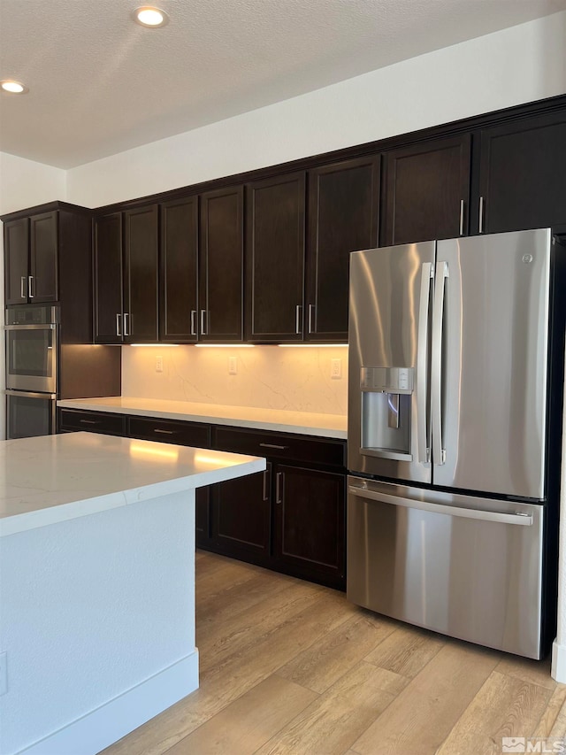 kitchen with a textured ceiling, light stone countertops, dark brown cabinets, light hardwood / wood-style floors, and stainless steel appliances