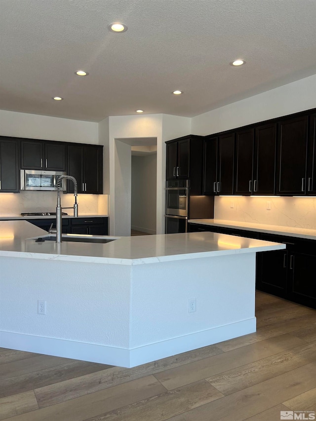 kitchen with a textured ceiling, appliances with stainless steel finishes, an island with sink, sink, and light wood-type flooring