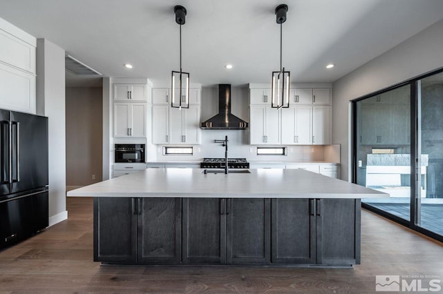 kitchen with white cabinets, decorative light fixtures, a large island with sink, wall chimney range hood, and black appliances