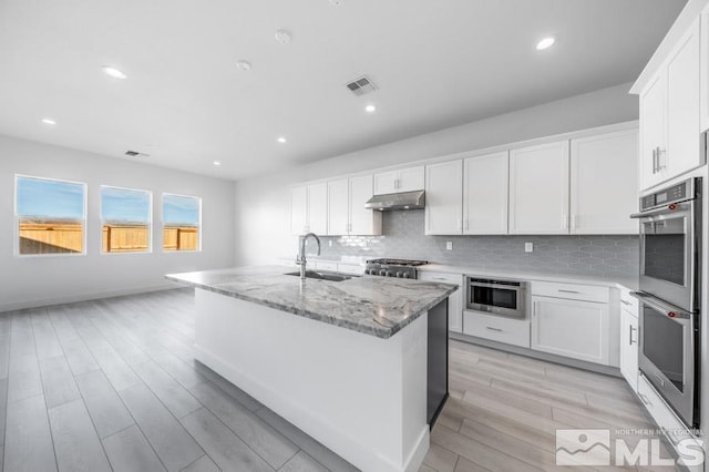 kitchen featuring light stone counters, double oven, sink, white cabinetry, and an island with sink