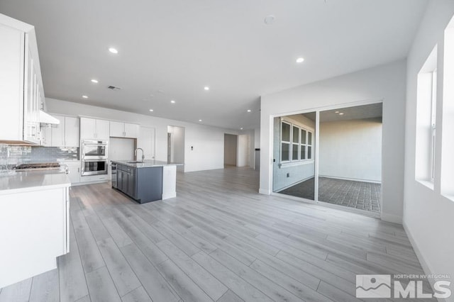 kitchen featuring a center island with sink, white cabinets, sink, decorative backsplash, and double oven