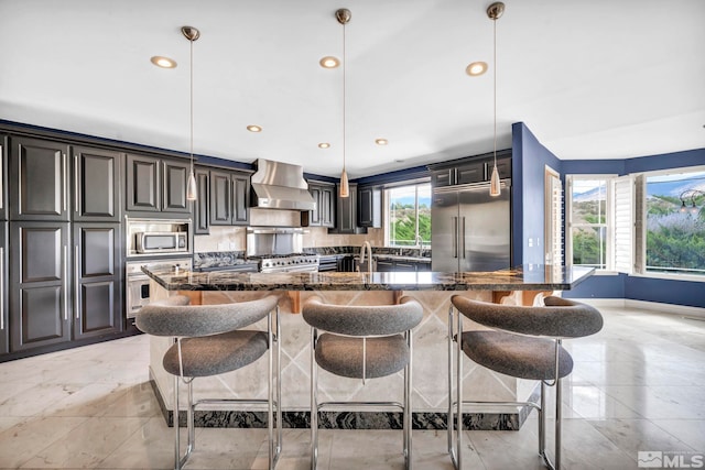 kitchen featuring a kitchen island, decorative light fixtures, wall chimney range hood, built in appliances, and dark stone countertops