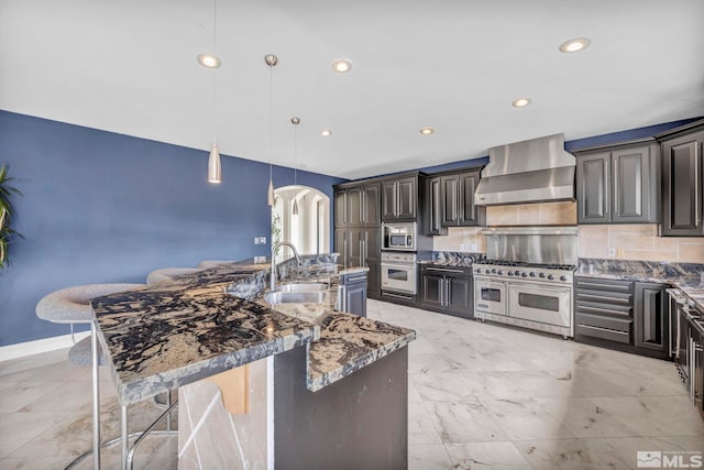 kitchen featuring decorative light fixtures, a large island with sink, wall chimney exhaust hood, and stainless steel appliances