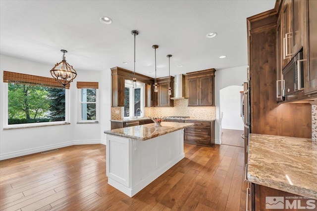 kitchen with light stone counters, decorative light fixtures, a kitchen island, and wall chimney range hood