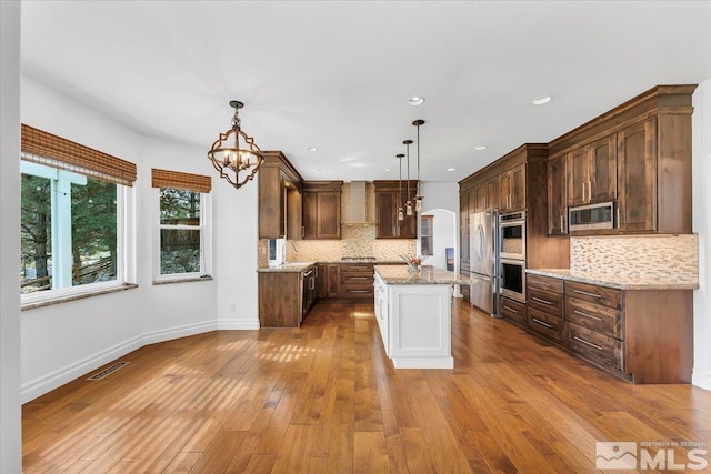 kitchen with appliances with stainless steel finishes, a kitchen island, light stone countertops, pendant lighting, and light hardwood / wood-style flooring