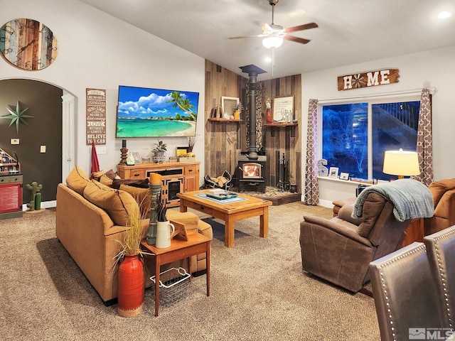 carpeted living room featuring ceiling fan, wooden walls, vaulted ceiling, and a wood stove