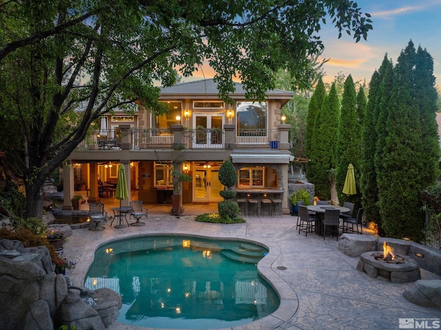 back house at dusk featuring a balcony, a patio area, a bar, and a fire pit