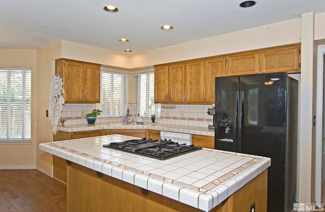 kitchen with backsplash, dark wood-type flooring, black fridge with ice dispenser, stainless steel gas stovetop, and tile countertops