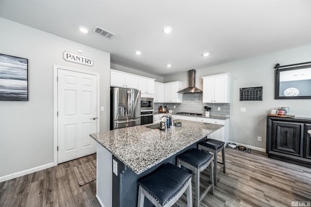 kitchen with white cabinetry, light stone countertops, stainless steel appliances, a center island with sink, and wall chimney range hood