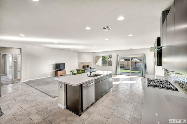 kitchen featuring a stone fireplace, dishwasher, light tile patterned floors, a center island with sink, and sink