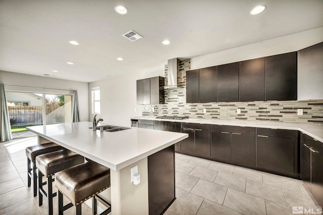 kitchen with sink, an island with sink, a breakfast bar area, stainless steel gas cooktop, and wall chimney exhaust hood