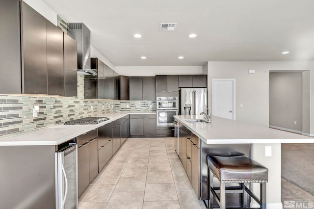 kitchen with a breakfast bar, an island with sink, wall chimney range hood, stainless steel appliances, and backsplash