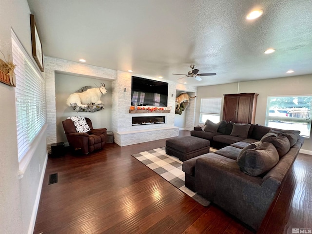 living room with a textured ceiling, a stone fireplace, dark hardwood / wood-style flooring, and ceiling fan