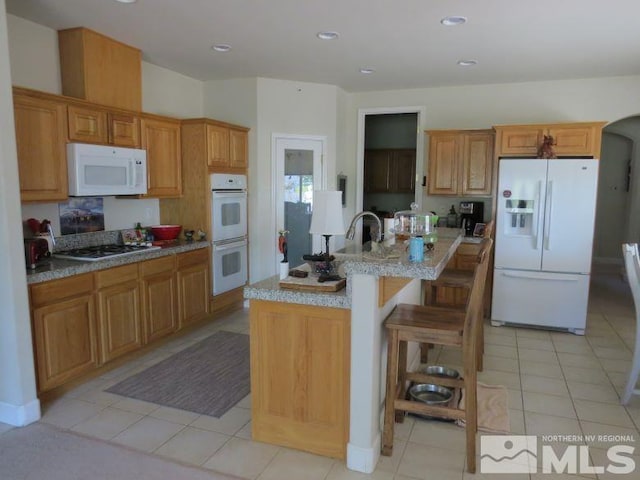 kitchen featuring light stone counters, light tile patterned flooring, white appliances, a center island with sink, and a breakfast bar