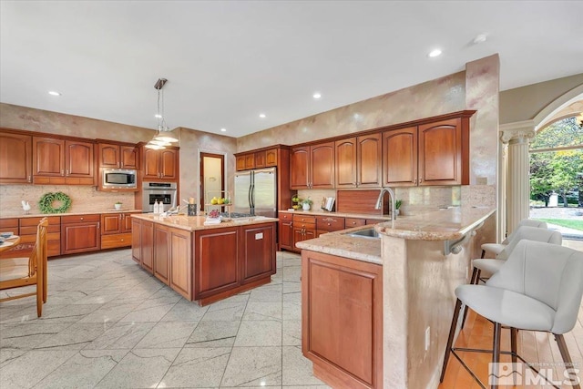 kitchen featuring a kitchen island with sink, sink, hanging light fixtures, appliances with stainless steel finishes, and ornate columns