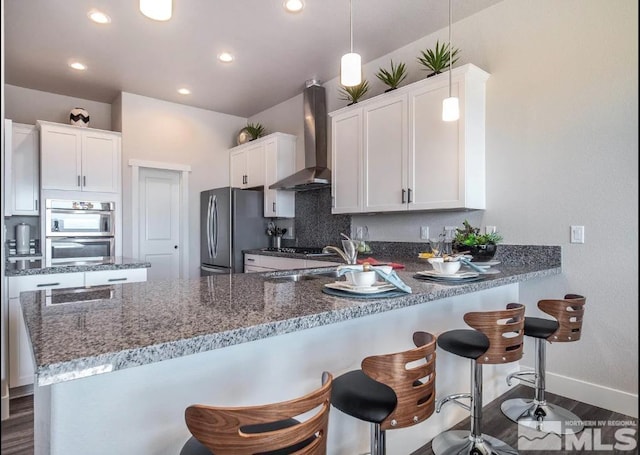 kitchen featuring appliances with stainless steel finishes, kitchen peninsula, wall chimney range hood, and white cabinetry