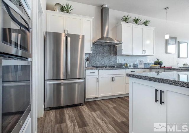 kitchen featuring appliances with stainless steel finishes, hanging light fixtures, white cabinetry, dark wood-type flooring, and wall chimney range hood