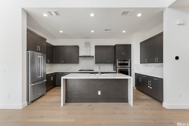 kitchen with appliances with stainless steel finishes, light hardwood / wood-style floors, dark brown cabinets, and an island with sink