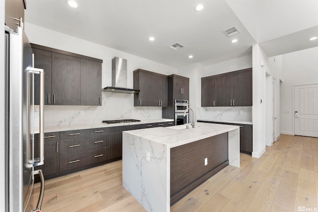 kitchen featuring a kitchen island with sink, wall chimney exhaust hood, light hardwood / wood-style flooring, appliances with stainless steel finishes, and light stone countertops