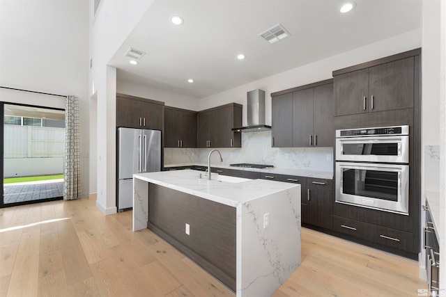 kitchen featuring light wood-type flooring, sink, wall chimney exhaust hood, a center island with sink, and appliances with stainless steel finishes