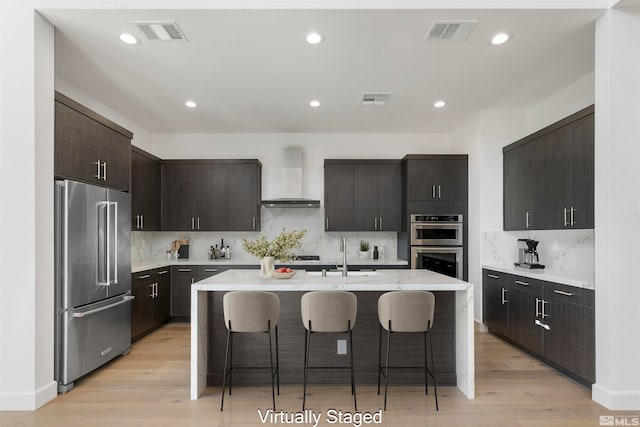 kitchen featuring a kitchen island with sink, wall chimney exhaust hood, stainless steel appliances, a kitchen breakfast bar, and light hardwood / wood-style flooring