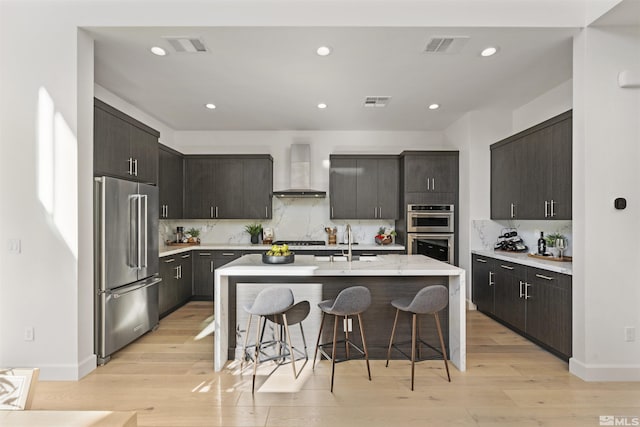 kitchen featuring light wood finished floors, wall chimney exhaust hood, stainless steel appliances, a sink, and a breakfast bar area