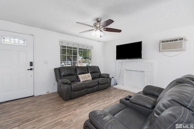 living room with a brick fireplace, ceiling fan, wood-type flooring, a wall mounted AC, and a textured ceiling