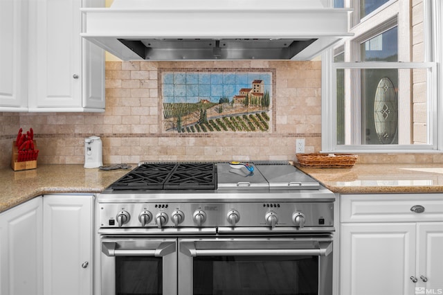 kitchen featuring high end stove, tasteful backsplash, light stone counters, premium range hood, and white cabinetry