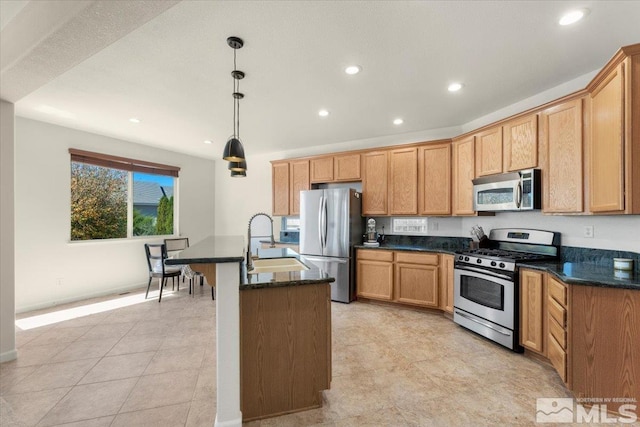 kitchen featuring a kitchen island with sink, dark stone counters, sink, stainless steel appliances, and hanging light fixtures