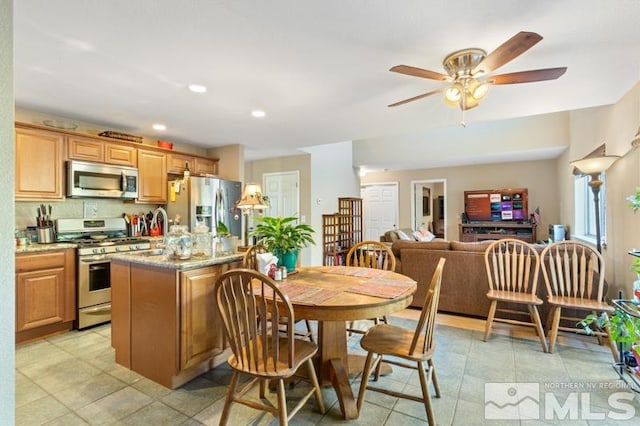 kitchen featuring tasteful backsplash, ceiling fan, stainless steel appliances, light stone counters, and a center island with sink