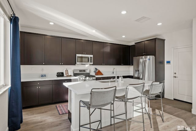 kitchen with dark brown cabinets, a center island with sink, appliances with stainless steel finishes, a breakfast bar, and light wood-type flooring