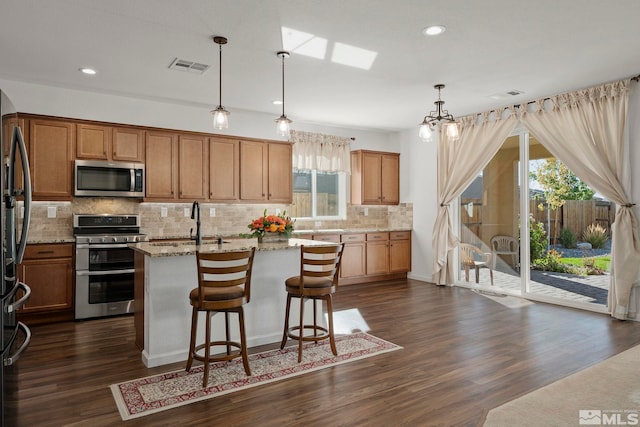 kitchen with light stone countertops, an island with sink, dark hardwood / wood-style flooring, stainless steel appliances, and pendant lighting