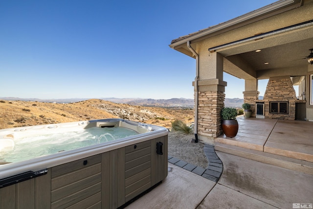 view of patio featuring a hot tub, an outdoor stone fireplace, a mountain view, and ceiling fan