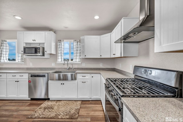 kitchen with appliances with stainless steel finishes, sink, light wood-type flooring, wall chimney exhaust hood, and white cabinets