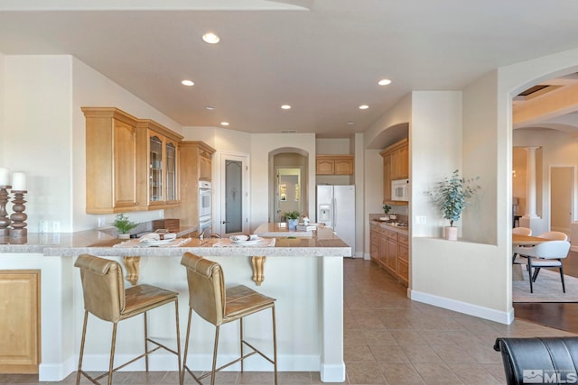 kitchen featuring white appliances, light brown cabinetry, a breakfast bar, kitchen peninsula, and light tile patterned floors