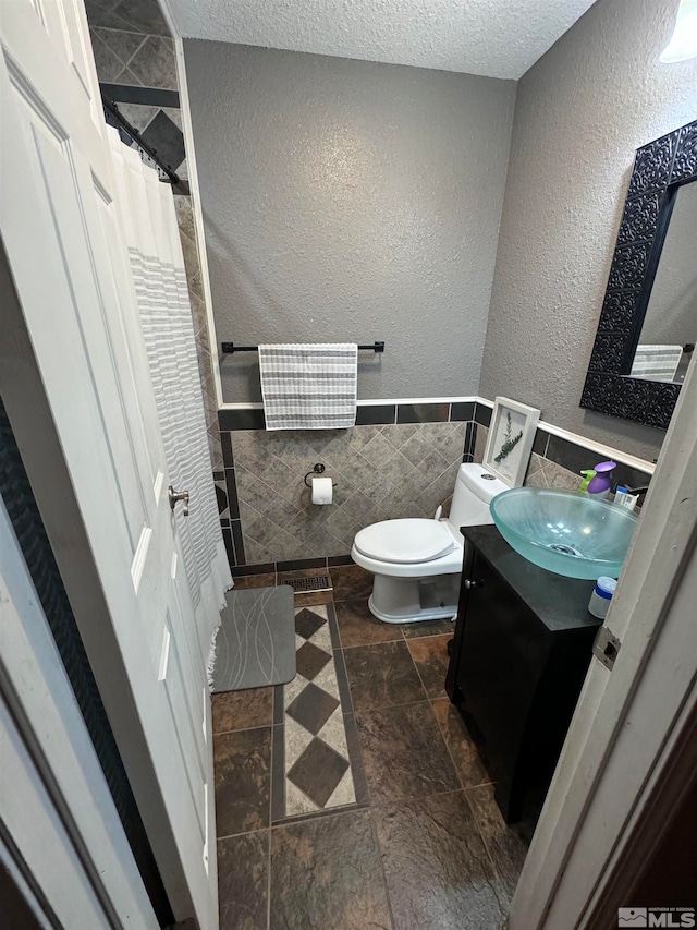 bathroom with a textured ceiling, toilet, walk in shower, tile walls, and vanity