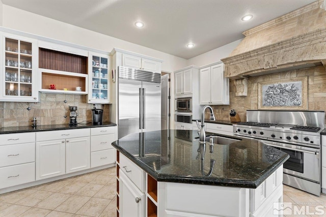 kitchen featuring sink, backsplash, built in appliances, dark stone countertops, and a center island with sink