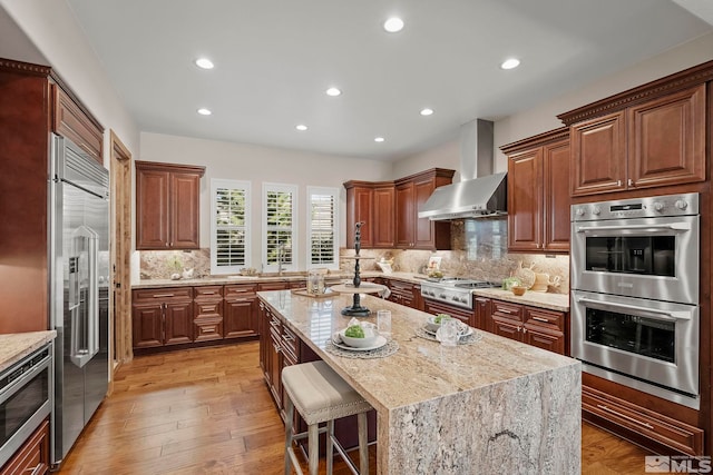 kitchen with wall chimney range hood, light stone counters, a breakfast bar area, light hardwood / wood-style flooring, and stainless steel appliances