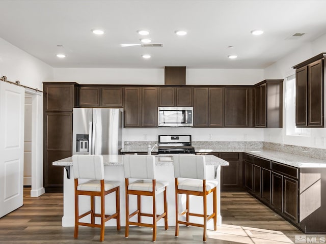 kitchen with appliances with stainless steel finishes, light stone countertops, a barn door, a kitchen island with sink, and dark wood-type flooring