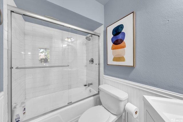 bathroom featuring bath / shower combo with glass door, a textured wall, toilet, and a wainscoted wall