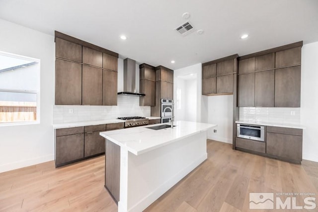 kitchen with a kitchen island with sink, sink, wall chimney exhaust hood, light wood-type flooring, and stainless steel appliances