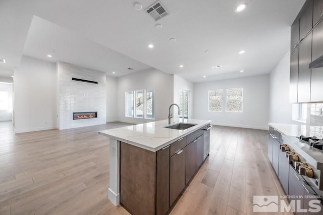 kitchen featuring a large fireplace, a kitchen island with sink, sink, and light wood-type flooring