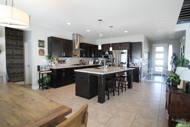 kitchen with wall chimney range hood, decorative backsplash, a kitchen island with sink, pendant lighting, and stainless steel appliances