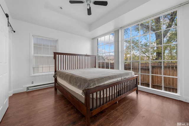 bedroom with ceiling fan, a barn door, a tray ceiling, dark wood-type flooring, and baseboard heating