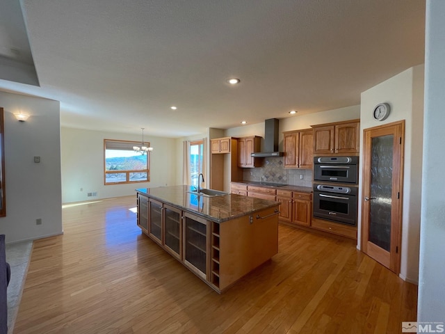 kitchen featuring wall chimney range hood, light hardwood / wood-style flooring, decorative backsplash, and an island with sink